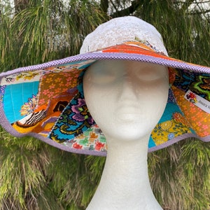 Puede incluir: Un sombrero de sol de ala ancha con un diseño de patchwork que presenta varios colores y patrones. El sombrero tiene una parte superior de malla blanca y un ribete morado. La etiqueta "Jessica Kolf" es visible en el sombrero.