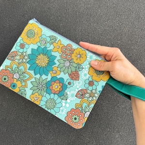 May include: A floral print clutch bag with a teal background and various flowers in yellow, orange, and teal. It features a teal zip and a teal wrist strap.