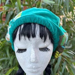 May include: A teal green beret hat with patterned fabric details. The hat is displayed on a mannequin head with black braids and false eyelashes. The background features blurred green foliage.