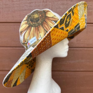 May include: A wide-brimmed sun hat with a patchwork design featuring sunflowers, geometric patterns, and orange, yellow, and brown colors. The hat is made of fabric and has a white label with the text "Sun Hat" on it.