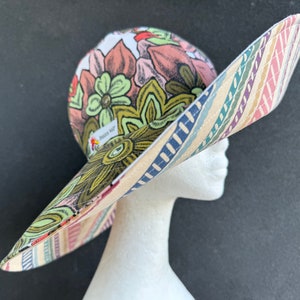 May include: A wide-brimmed sun hat with a floral print and a striped pattern on the brim. The hat is made of fabric and has a large, floppy brim. The hat is shown on a white mannequin head.