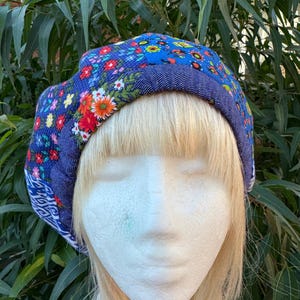 Puede incluir: Boina azul con estampado floral y ribete de mezclilla. La boina presenta una vibrante mezcla de flores rojas, naranjas, amarillas y blancas sobre un fondo azul. El borde es de mezclilla azul más oscura.