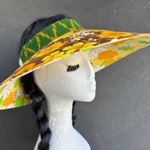 May include: A vintage-style visor with a wide brim, featuring a vibrant floral pattern in yellow, brown, and white. The headband is green with a geometric design. The visor is displayed on a mannequin head with black hair.