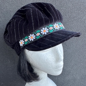 Op de afbeelding: Een zwarte baret met witte krijtstrepen en een decoratieve band. De band heeft een bloemmotief met witte madeliefjes, oranje centra en groene bladeren. De pet wordt getoond op een mannequinhoofd.
