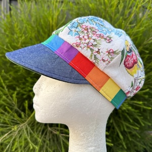 Könnte beinhalten: Eine weiße Leinenmütze mit Blumendruck und einem regenbogenfarbenen Band. Die Mütze hat ein Jeansvisier.
