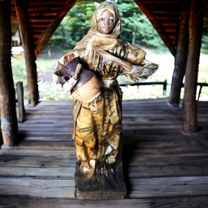 May include: A ceramic figurine of a woman wearing a brown and white robe, holding a brown clay pot. The figurine is standing on a wooden surface.