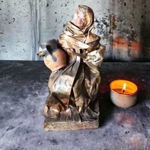 May include: A ceramic figurine of a person wearing a brown robe and holding a clay pot. The figurine is sitting on a wooden base. A small candle is lit in the background.