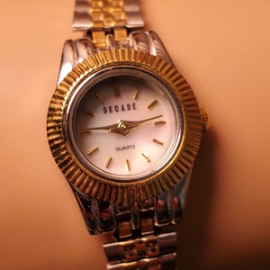 May include: A gold and silver two-tone watch with a white face and black hands. The watch face has the word "DECADE" and "QUARTZ" printed on it.