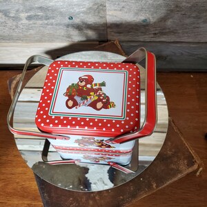 Puede incluir: Caja de lata roja y blanca con un patrón de lunares y un diseño de oso navideño. La caja tiene un asa y está apilada encima de otra caja similar.