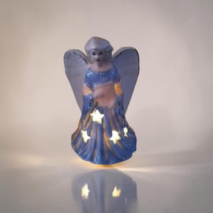 Könnte beinhalten: Eine Keramik-Engelsfigur in Blau und Weiß. Der Engel hat ausgebreitete Flügel und ein fließendes Gewand mit sternförmigen Aussparungen, die Licht emittieren. Ein dekorativer Artikel für die Inneneinrichtung.
