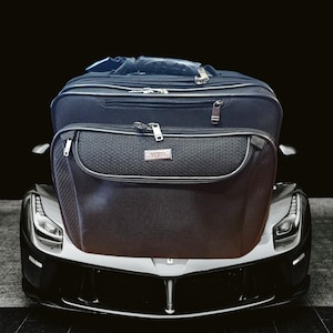 May include: A black travel bag sits on the front of a silver sports car. The bag has a zippered pocket and a black leather handle.