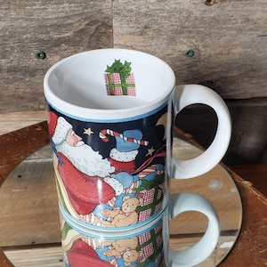 May include: White ceramic Christmas mug featuring a festive design of Santa Claus in a sleigh, with gifts and teddy bears. The mug has a white handle and a gift box graphic inside the rim. The background is a dark blue with stars and a crescent moon.