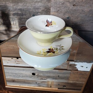 Puede incluir: Una taza de té amarilla y un platillo con un patrón floral con hojas marrones y flores amarillas. La taza y el platillo tienen un borde dorado.
