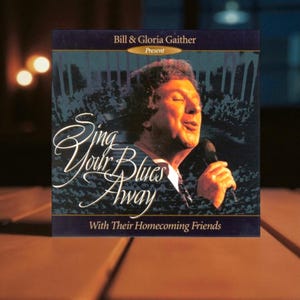 May include: A CD case featuring a man singing into a microphone. The text on the case reads "Sing Your Blues Away" and "With Their Homecoming Friends." The case is dark blue with a gold border and the words "Bill & Gloria Gaither Present."