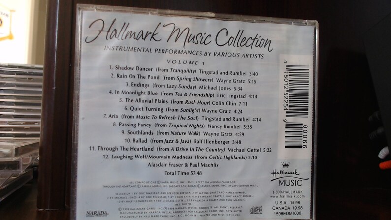 May include: The back of a CD case for the Hallmark Music Collection, Volume 1. The case is white with black text listing the 12 instrumental tracks on the CD, including the title, artist, and album from which the song originates. The case also includes the total time of the CD, 57:48, and the names of the producers, Alasdair Fraser and Paul Machlis. The case has a barcode and a Hallmark Music logo.