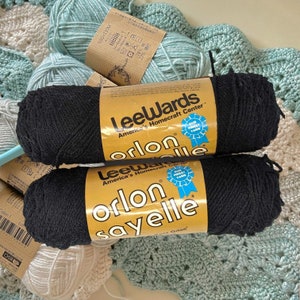May include: Two spools of black yarn labeled "LeeWards Orlon" and "LeeWards Orlon Sayelle". The yarn is packaged in yellow and white labels with red and blue accents. The labels have the text "Our Finest Yarn" printed on them.