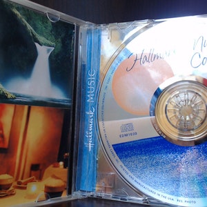 May include: A silver CD with a blue and white design. The CD is labeled "Hallmark Music Collection" and features a crown logo. The CD is in a clear plastic case with a photo of a waterfall on the front.