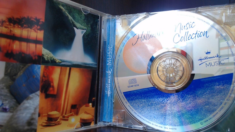May include: A silver CD with a blue and white design. The CD is labeled "Hallmark Music Collection" and features a crown logo. The CD is in a clear plastic case with a photo of a waterfall on the front.