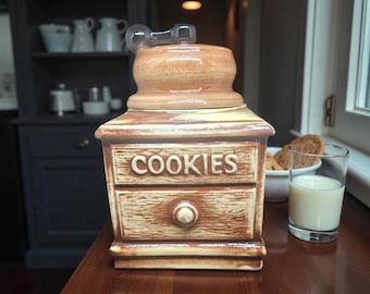 Könnte beinhalten: Ein Keramik-Keksdose im Vintage-Stil mit hellbraunem und beigem Finish. Die Dose hat die Form eines Schranks mit zwei Schubladen, und das Wort "COOKIES" steht oben. Ein Glas Milch und eine Schüssel mit Keksen befinden sich in der Nähe.