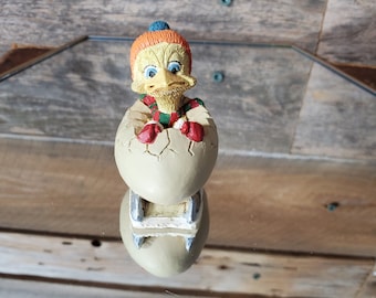 Peut inclure: Un oiseau jaune et orange portant un chapeau bleu et blanc et une écharpe rouge et verte est assis dans une coquille d'œuf fissurée. L'oiseau tient une moufle rouge dans chaque main. L'œuf est assis sur un traîneau en bois.