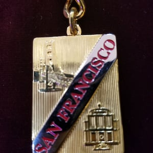 May include: Gold-toned metal keychain featuring a rectangular pendant with vertical lines. The pendant has an angled black and silver stripe with "SAN FRANCISCO" in red lettering. The pendant also features images of the Golden Gate Bridge and other landmarks.