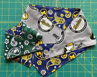 May include: Three dog bandanas in a layered arrangement. One is green with white floral and paisley patterns, another is blue with yellow paisley and floral patterns, and the third is grey with a university logo and text.