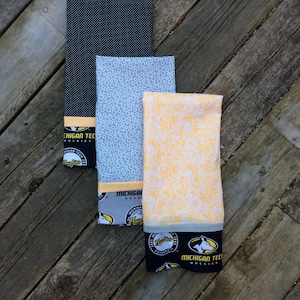 May include: Three kitchen towels with a black, white, and yellow color scheme. The towels feature a repeating pattern of circles, a floral design, and a Michigan Tech Huskies logo. The towels are made of cotton and are perfect for drying dishes, hands, and surfaces.