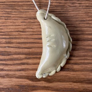 May include: A beige ceramic ornament shaped like a crescent moon with a rope attached to the top. The ornament has a textured edge and a small indentation on the surface.