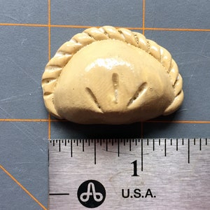 May include: A tan-colored, dumpling-shaped decorative item. The item has a braided edge and etched details on the front. The item is approximately 1 inch in length, as measured by the ruler in the image.