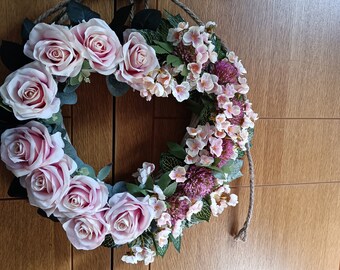 May include: A circular floral wreath featuring large, light pink roses and smaller pink and purple flowers. The wreath is accented with green leaves and is suspended by a rope loop. The wreath is a decorative item.