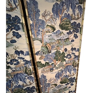 May include: Two-panel screen with a bamboo-style gold frame. The panels feature a scenic landscape design with blue, green, and beige tones. The artwork depicts trees, bridges, and figures in boats, creating a serene, traditional aesthetic.