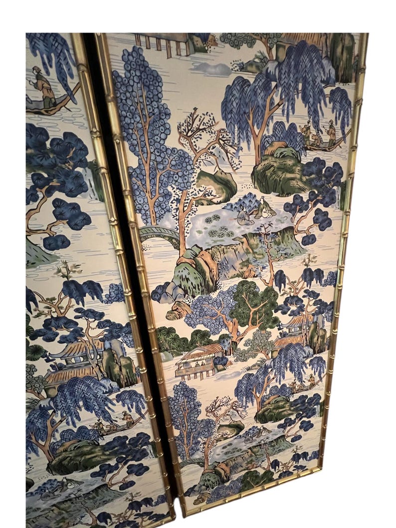 May include: Two-panel screen with a bamboo-style gold frame. The panels feature a scenic landscape design with blue, green, and beige tones. The artwork depicts trees, bridges, and figures in boats, creating a serene, traditional aesthetic.