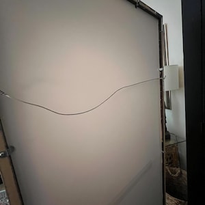 May include: Back of a large framed artwork with a light-colored surface. The frame is dark brown with a metal wire for hanging. The artwork is leaning against a wall, with a glimpse of a lamp and furniture in the background.