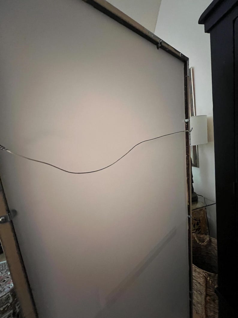 May include: Back of a large framed artwork with a light-colored surface. The frame is dark brown with a metal wire for hanging. The artwork is leaning against a wall, with a glimpse of a lamp and furniture in the background.