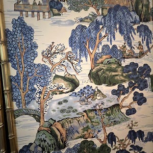 May include: A framed panel featuring a scenic landscape design. The artwork depicts a serene scene with blue and green trees, a bridge, and small buildings. The color palette includes shades of blue, green, and brown against a cream background. The frame is a gold bamboo style.