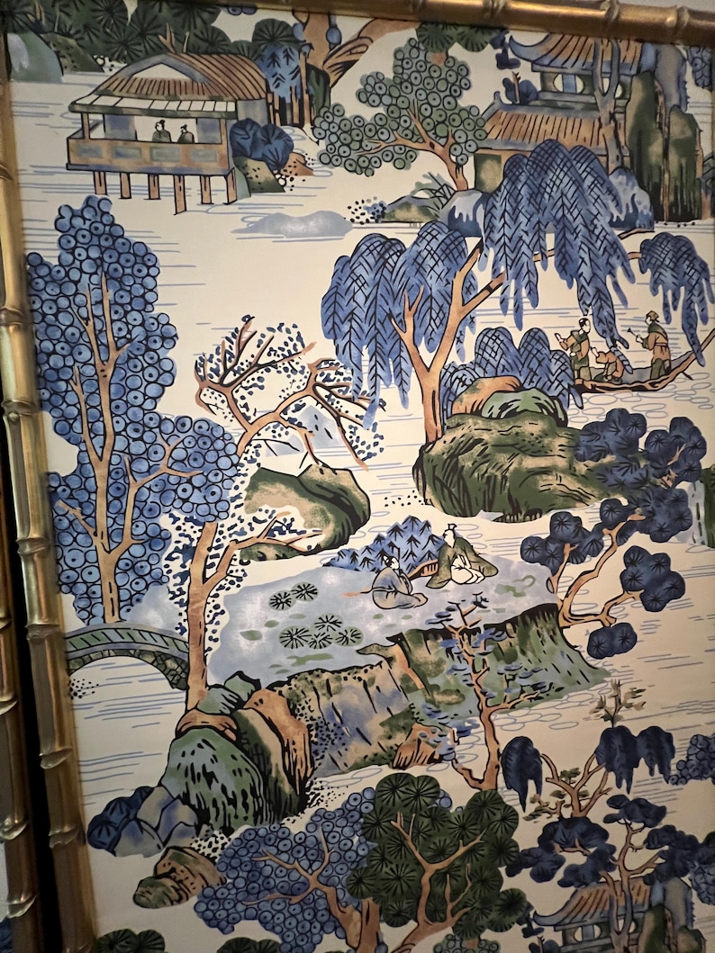 May include: A framed panel featuring a scenic landscape design. The artwork depicts a serene scene with blue and green trees, a bridge, and small buildings. The color palette includes shades of blue, green, and brown against a cream background. The frame is a gold bamboo style.