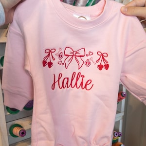 May include: A light pink baby romper with long sleeves. The romper features red embroidered designs of bows, hearts, keys, and cherries. The name "Hallie" is embroidered in red script below the design.