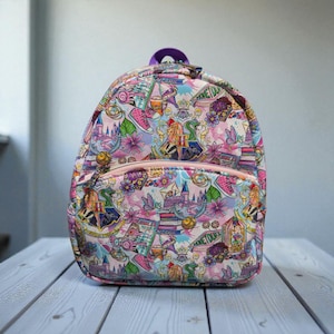 May include: A colorful backpack with a pink zipper and a purple handle. The backpack is covered in a repeating pattern of castles, flowers, and other fantasy-themed illustrations in shades of pink, purple, and blue.
