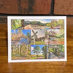 May include: A white-bordered postcard featuring a collage of scenic bridge and railroad crossing images. The images showcase various bridges, a railroad crossing sign, and natural landscapes with trees and water. The postcard is displayed on a wooden surface against a brick background.