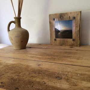 Pu&ograve; includere: Una cornice per foto in legno rustico con un inserto in acrilico trasparente, che mostra una fotografia di una scena di spiaggia. La cornice &egrave; appoggiata su un tavolo in legno con un'anfora di terracotta marrone.