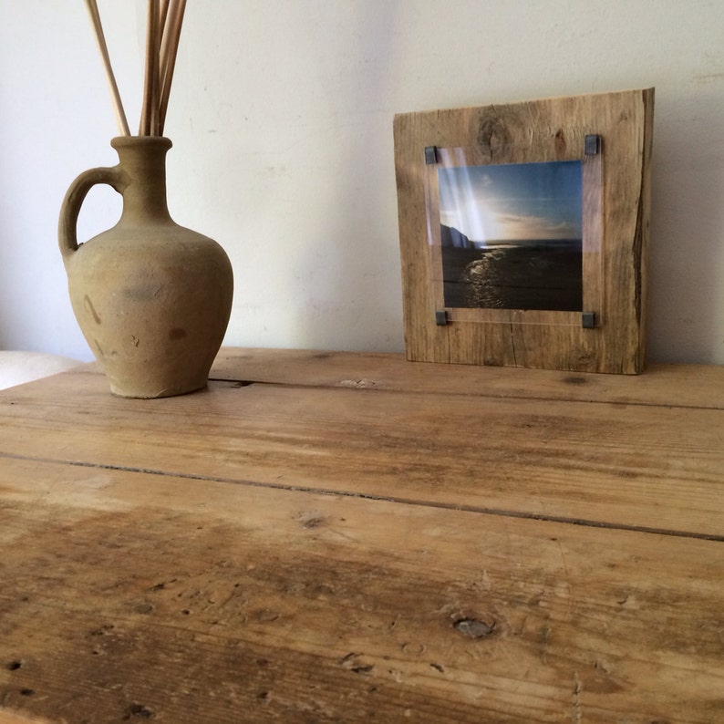 Pu&ograve; includere: Una cornice per foto in legno rustico con un inserto in acrilico trasparente, che mostra una fotografia di una scena di spiaggia. La cornice &egrave; appoggiata su un tavolo in legno con un'anfora di terracotta marrone.