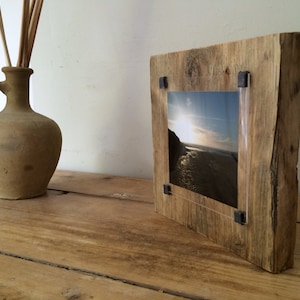 Pu&ograve; includere: Cornice per foto in legno rustico con finitura in legno naturale e inserto in acrilico trasparente. La cornice contiene una fotografia di una scena di spiaggia con un tramonto.