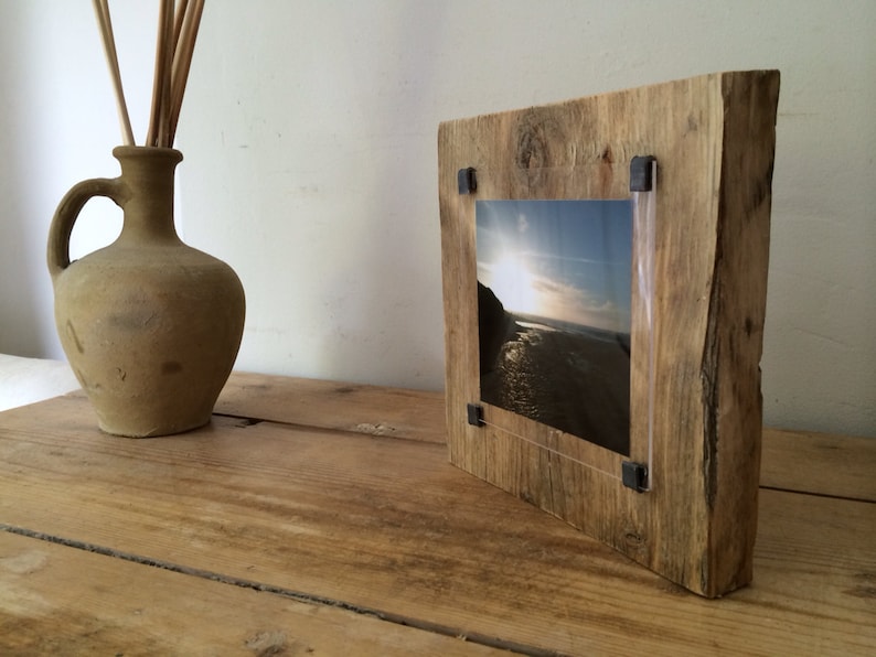 Pu&ograve; includere: Cornice per foto in legno rustico con finitura in legno naturale e inserto in acrilico trasparente. La cornice contiene una fotografia di una scena di spiaggia con un tramonto.
