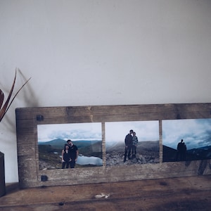 Może przedstawiać: Rustykalna drewniana ramka na zdjęcia z trzema fotografiami krajobrazowymi. Ramka wykonana jest z postarzanego drewna i ma metalowe akcenty. Zdjęcia przedstawiają pary w górskich sceneriach, z jeziorem w jednym.