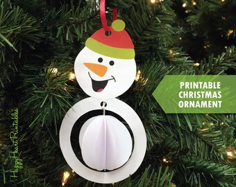 May include: A white snowman Christmas ornament with a red hat and green brim, hanging from a red ribbon. The ornament has a 3D paper ball body and a smiling face. Text on the image reads "Printable Christmas Ornament."