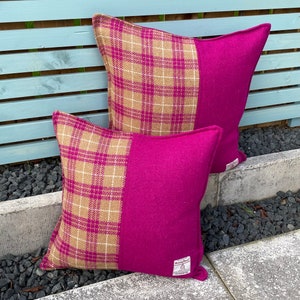 Op de afbeelding: Twee vierkante kussenslopen met een geruit patroon aan de ene kant en een effen magenta kleur aan de andere kant. De kussens zijn gemaakt van een zachte, textuurrijke stof. De kussens liggen op een stenen oppervlak.