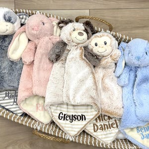 May include: Five plush animal security blankets in a woven basket. The collection includes a gray cow, a pink rabbit, a brown and white bear, a monkey, and a blue rabbit. Each blanket features embroidered names including Greyson, Daniel, and Bennett James.