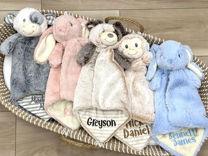 May include: Five plush animal security blankets in a woven basket. The collection includes a gray cow, a pink rabbit, a brown and white bear, a monkey, and a blue rabbit. Each blanket features embroidered names including Greyson, Daniel, and Bennett James.