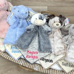 May include: Five plush animal security blankets with embroidered names in a woven basket. The collection includes a pink bunny, blue bunny, gray and white cow, brown and white puppy, and a gray bunny, each featuring soft fabric and personalized text.