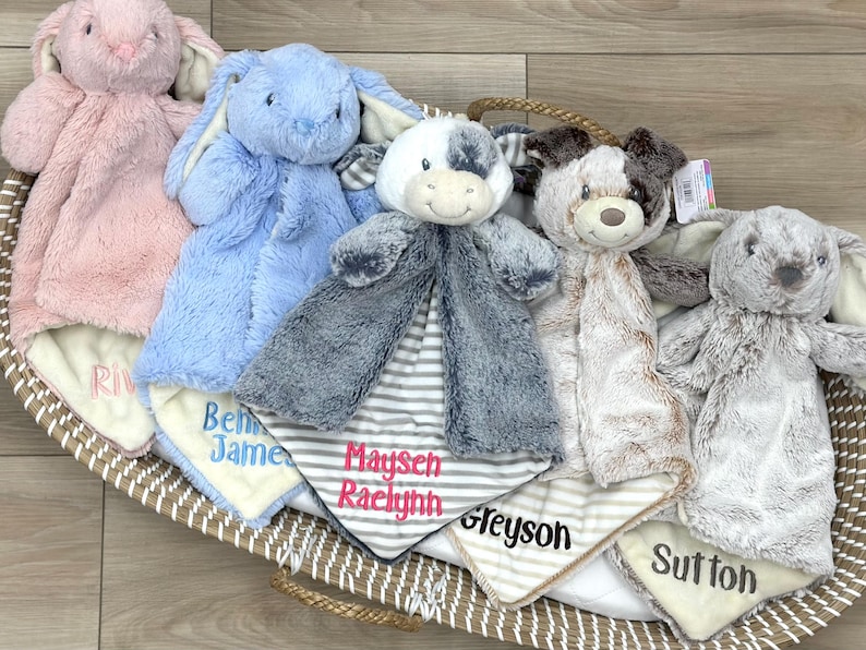 May include: Five plush animal security blankets with embroidered names in a woven basket. The collection includes a pink bunny, blue bunny, gray and white cow, brown and white puppy, and a gray bunny, each featuring soft fabric and personalized text.